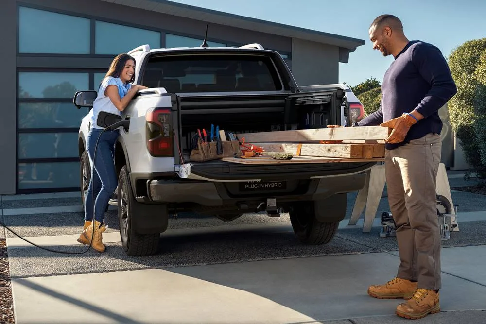 A white Ford Ranger Plug-in Hybrid ute is charging in a driveway while a man loads timber.