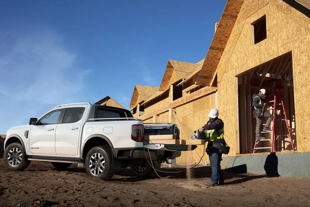A white Ford Ranger Wildtrak ute on a construction site, with a worker using a power saw plugged into the open tailgate.