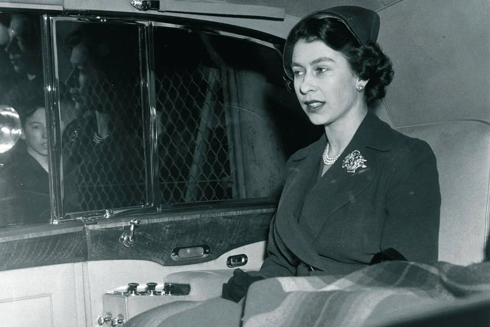 A black and white photo of a young Queen Elizabeth II in a luxurious car, wearing a hat, pearls and a brooch.
