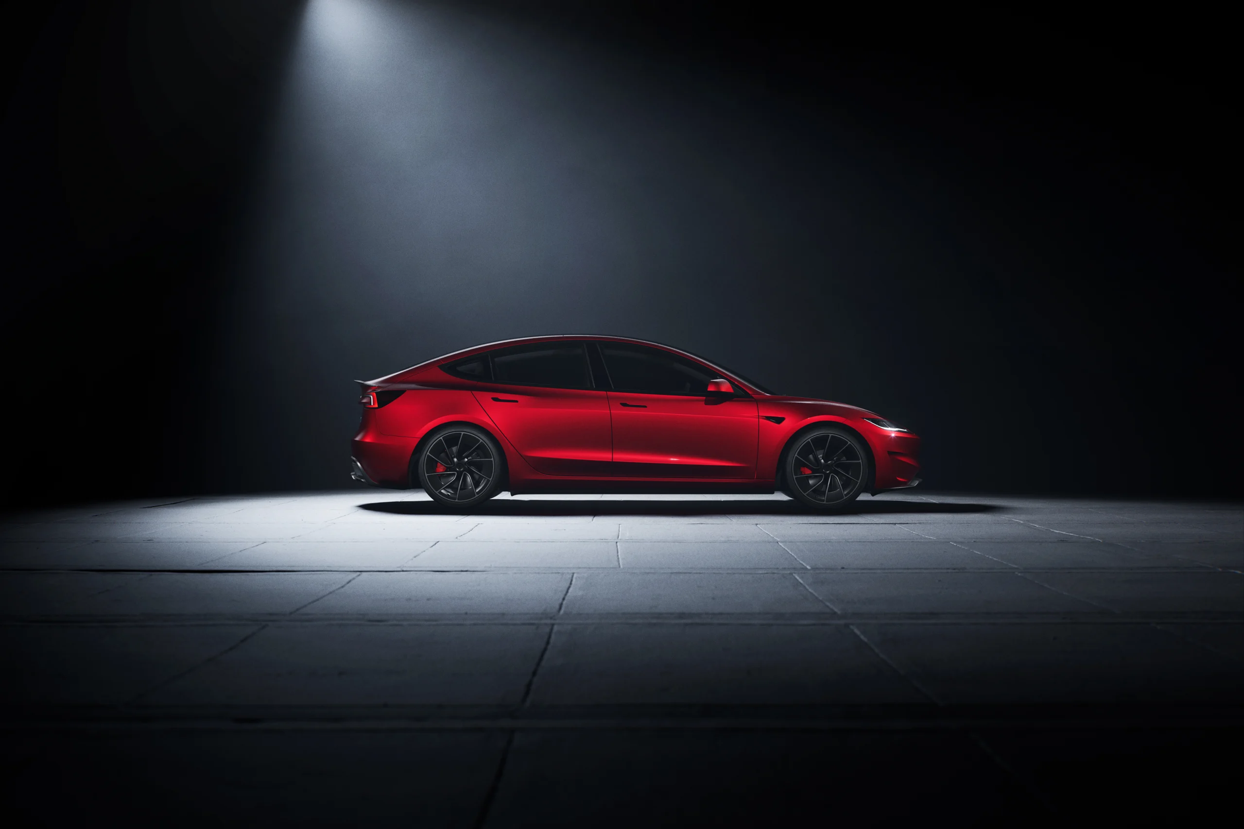 Red Tesla Model 3 Performance sedan in side profile, illuminated by a single spotlight on a dark, textured floor.