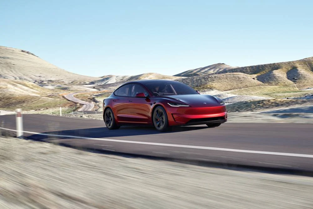 Deep red Tesla Model 3 Performance driving on a winding road through arid, hilly terrain under a clear blue sky.