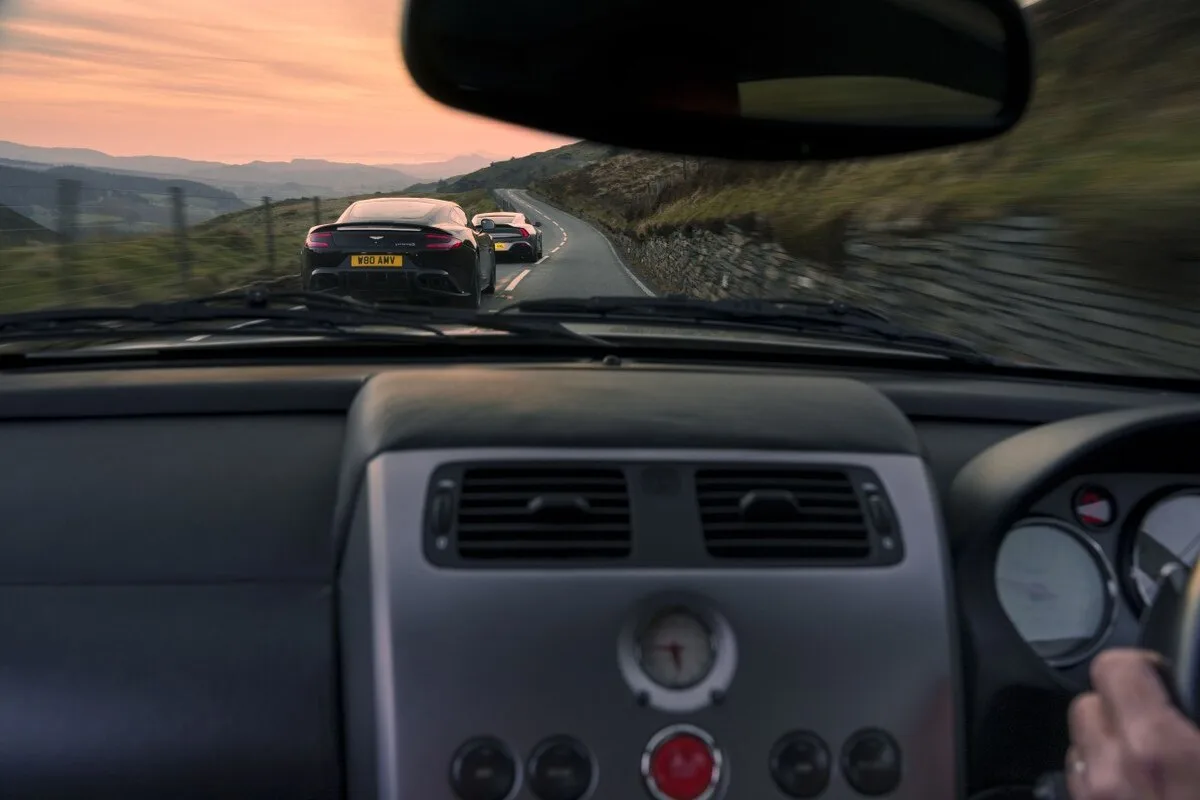 View from inside original Aston Martin Vanquish following two later generations on mountain road at sunset