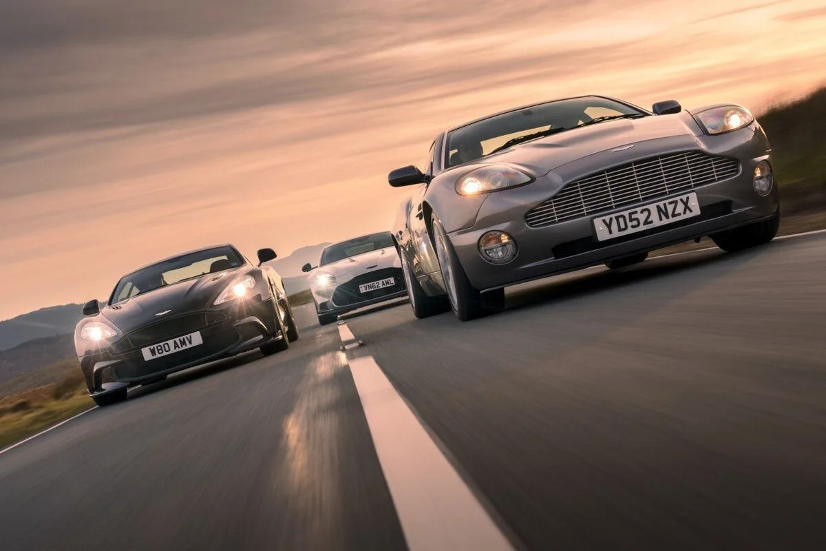 Three generations of Aston Martin Vanquish in low angle dynamic shot at sunset with original 2001 model leading