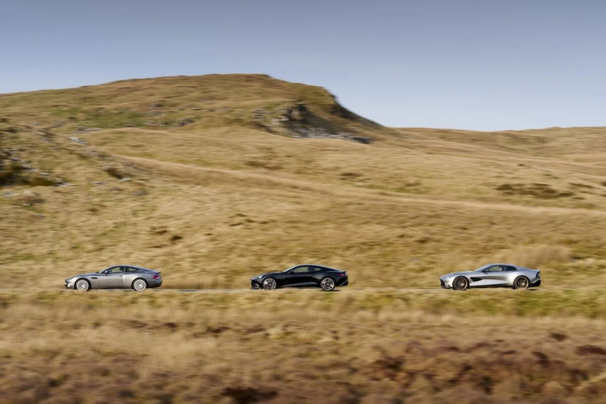Three Aston Martin Vanquish generations in profile against Welsh hills backdrop