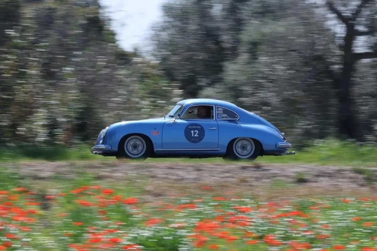 Side view of a light blue vintage Porsche 356 rally coupe with number 12 driving past poppies and trees.