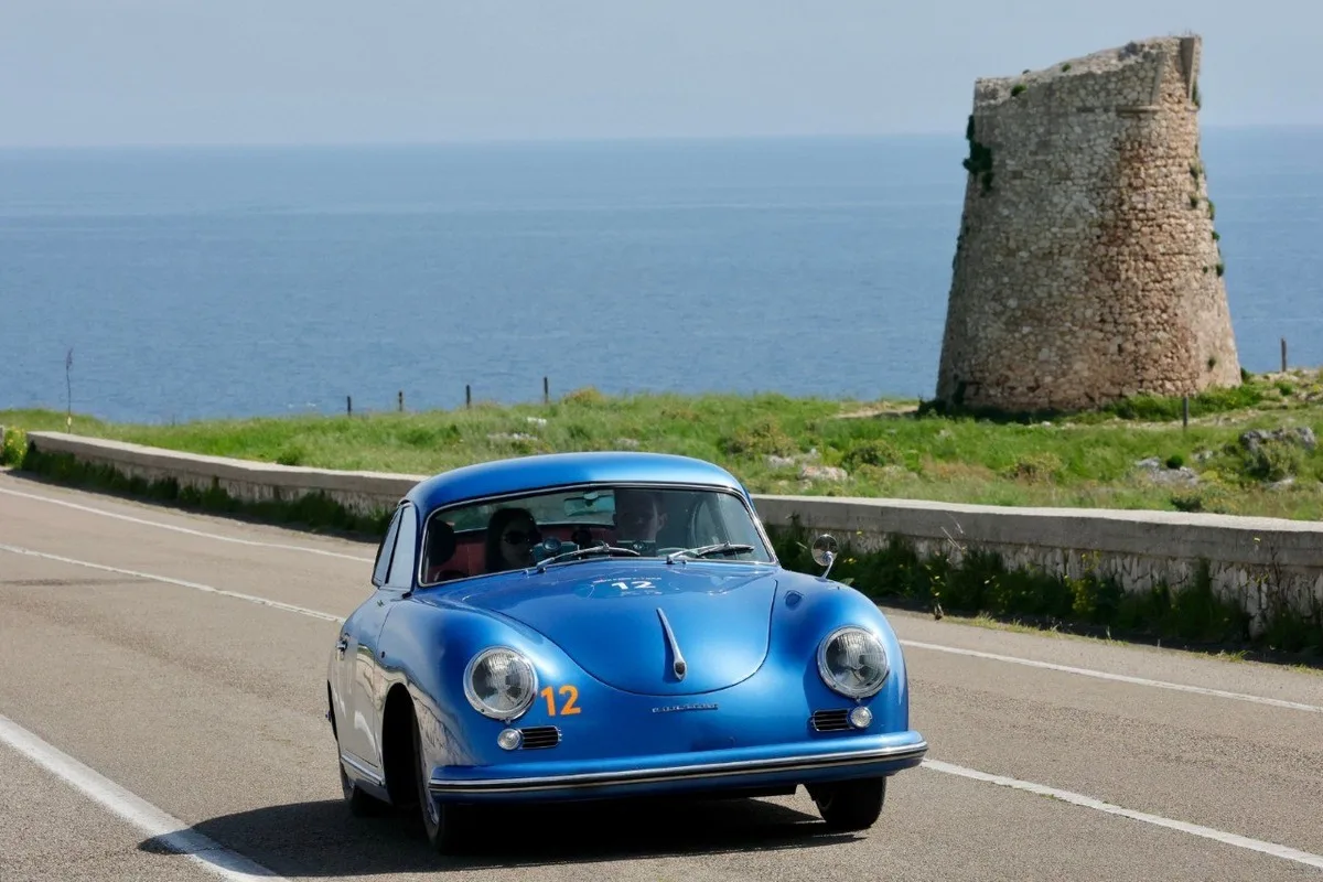 Front view of a blue Porsche 356 rally car number 12 on a seaside road with the ocean and a stone tower behind.