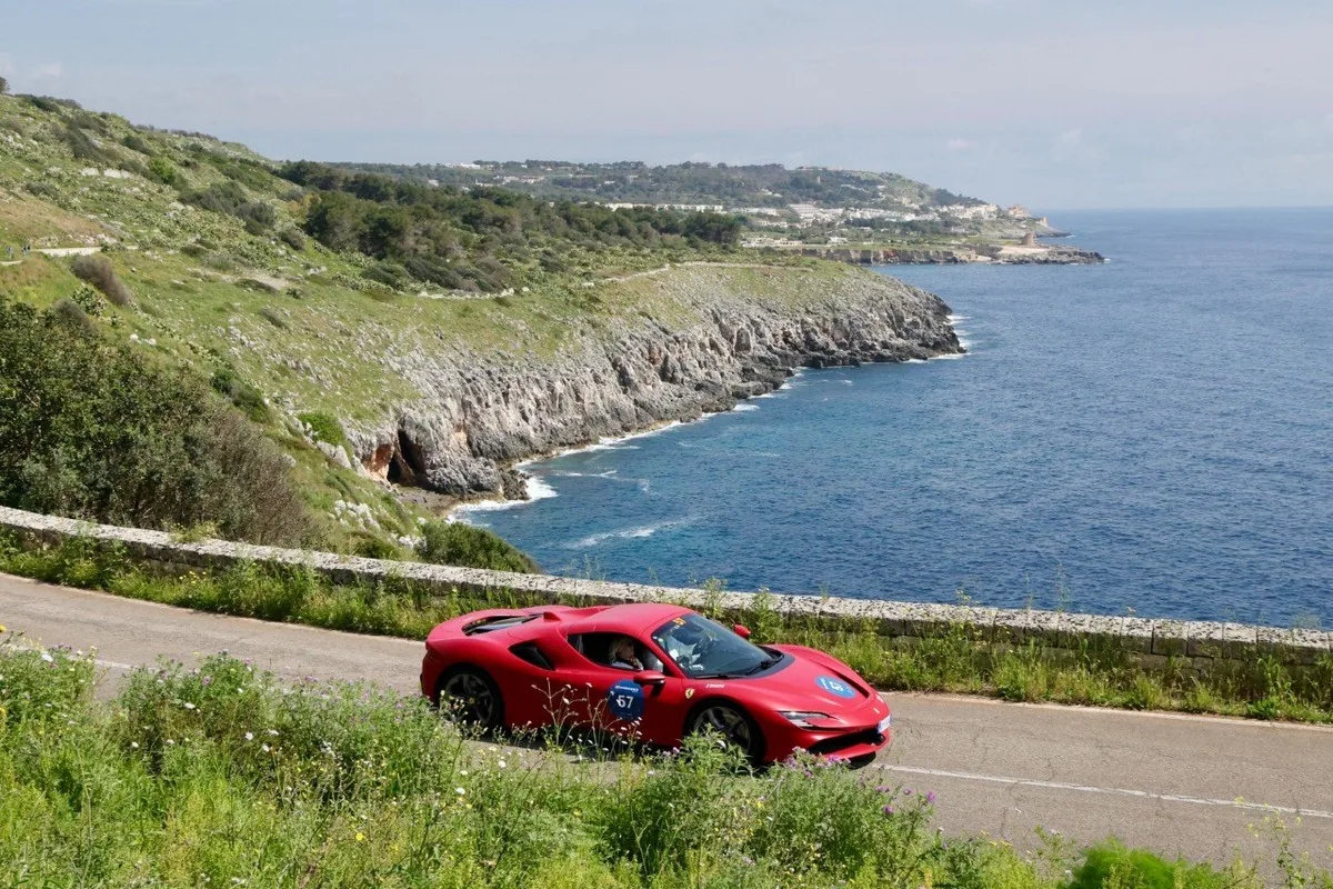 Red Ferrari 296 GTB coupe with rally number 67 driving along a scenic coastal road above rocky seaside cliffs.