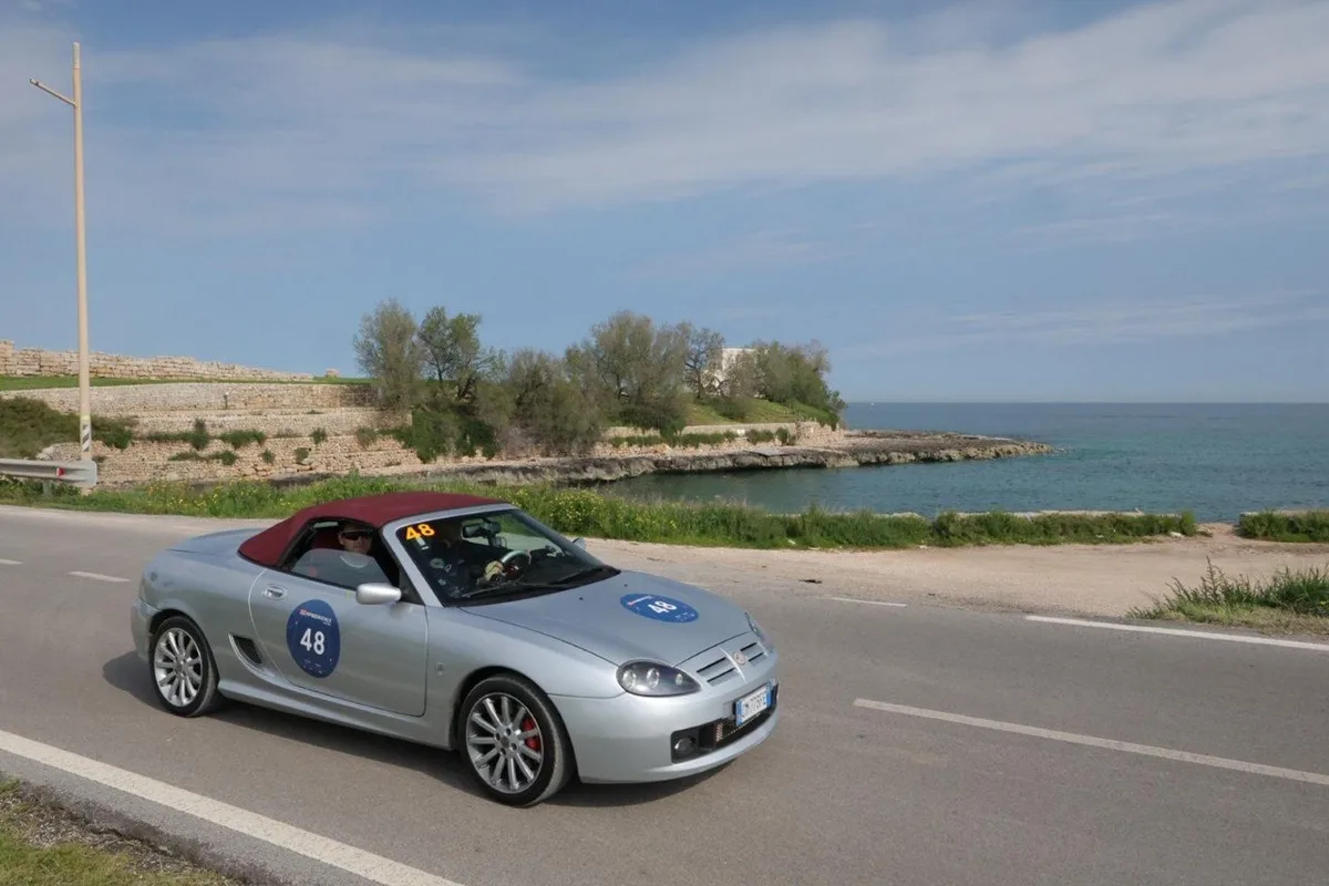 Silver MG TF convertible with burgundy soft top and rally number 48 on a coastal road by the sea.
