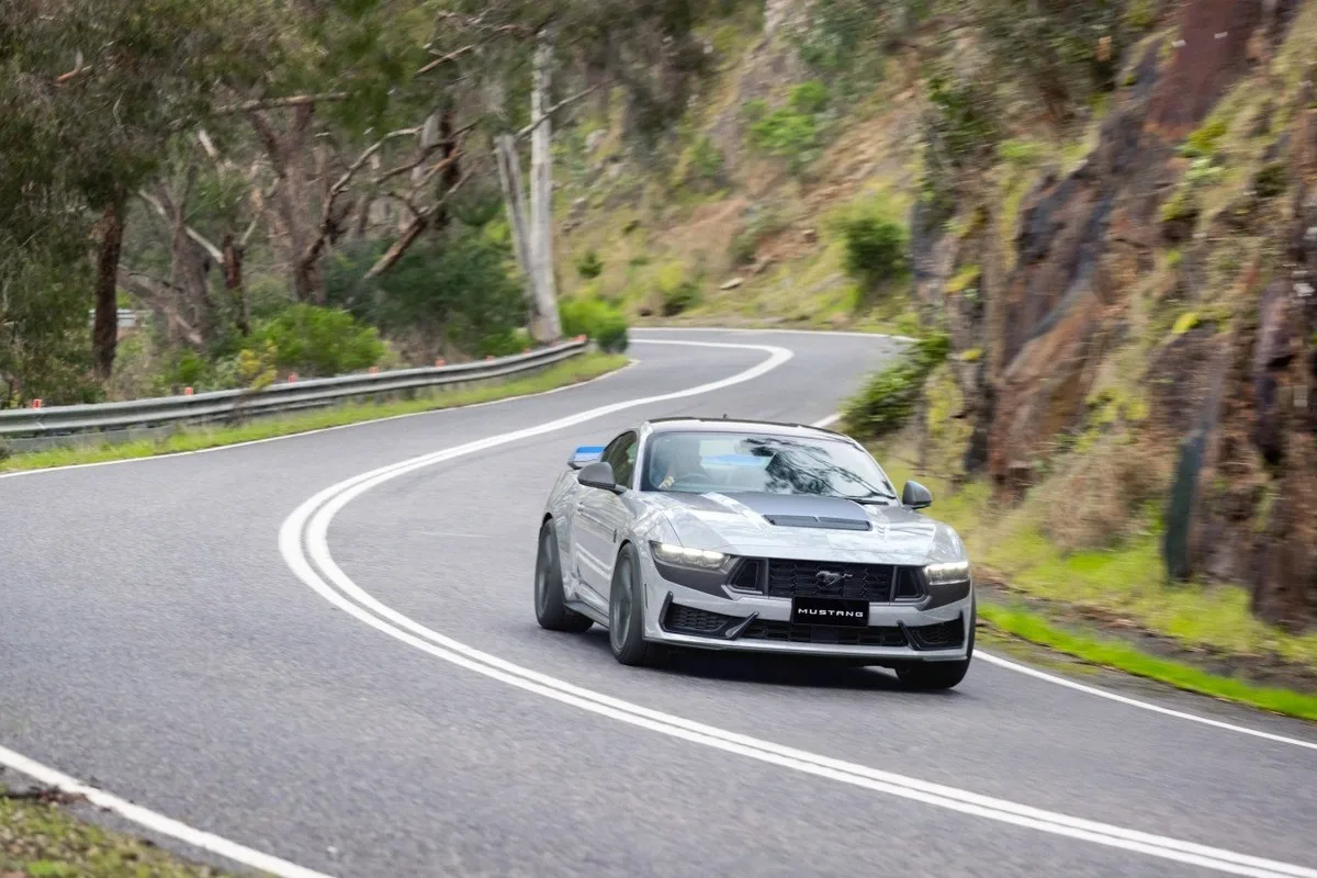 Silver Ford Mustang Dark Horse rounds a forest road with headlights on.