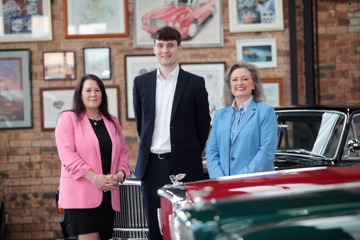 Three people pose in a classic car showroom with vintage luxury cars and framed automotive art behind them.