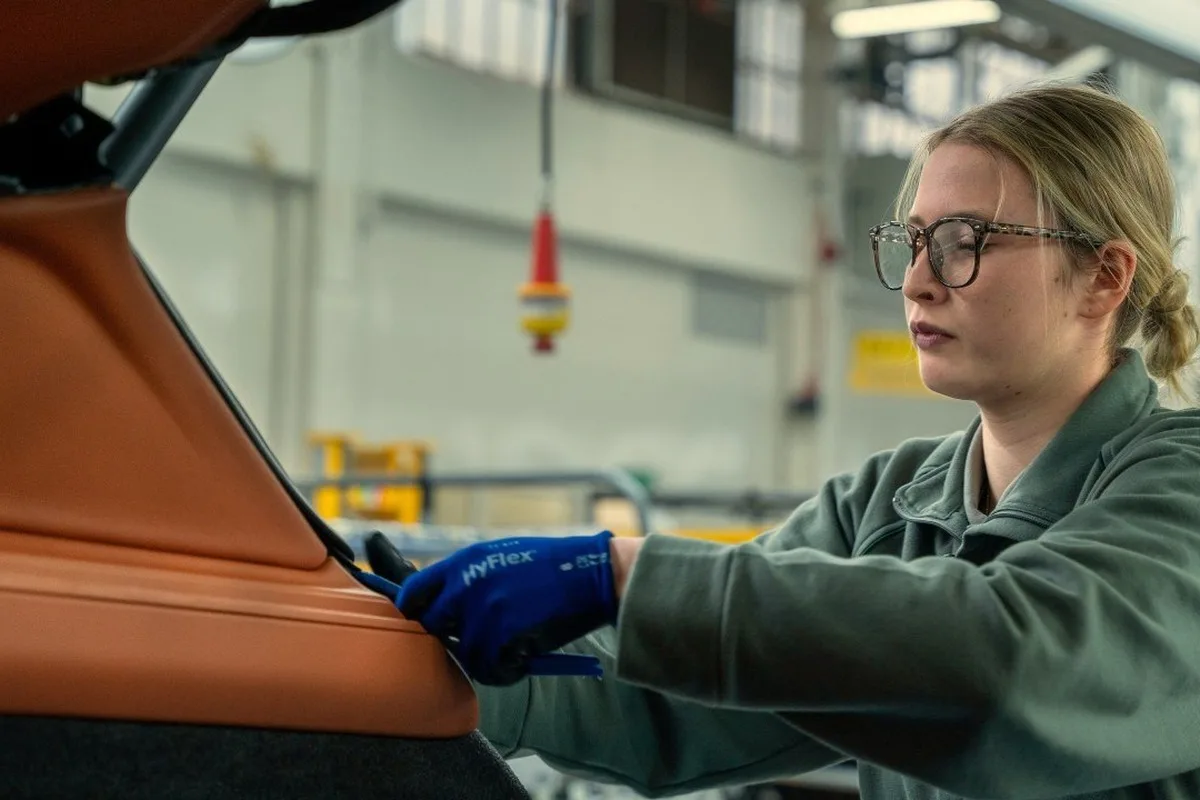 A factory worker in gloves and protective glasses works on an orange vehicle component on the assembly line.
