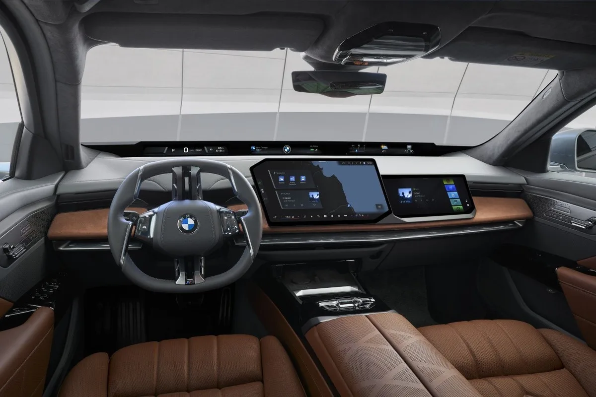 Front interior of a modern BMW showing steering wheel, panoramic digital display, central touchscreen, passenger display, wood trim, and brown leather seats.