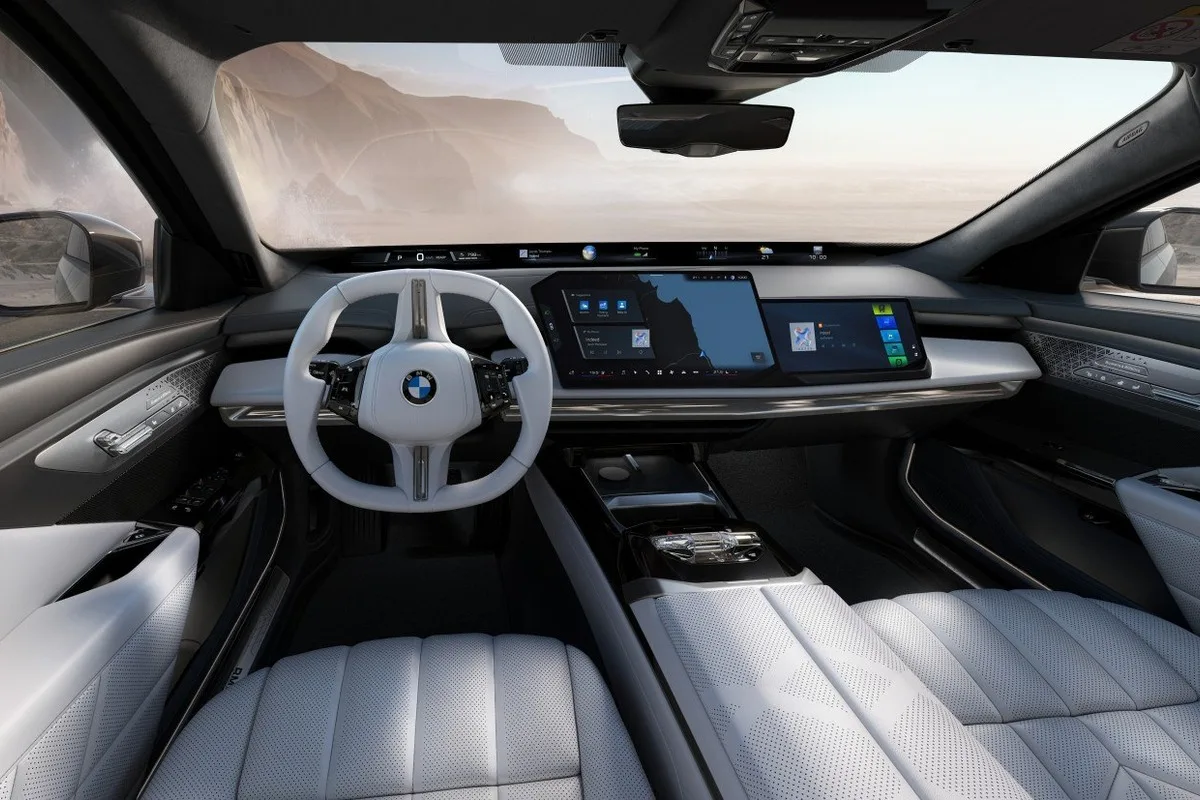 Front interior of a BMW luxury car with white leather seats, white steering wheel, wide digital dashboard screens, and centre console viewed from the back seat.
