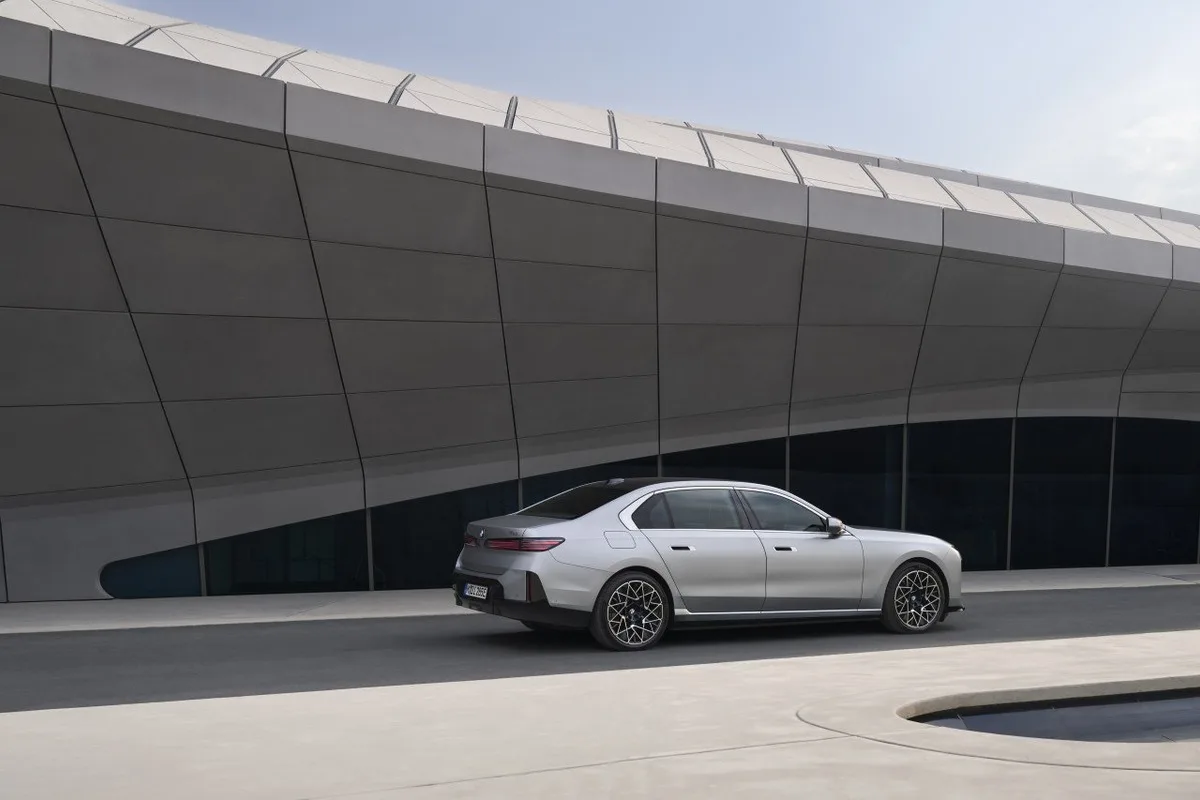 Silver four-door luxury sedan parked on a paved road beside a large curved modern building with dark metallic panels.