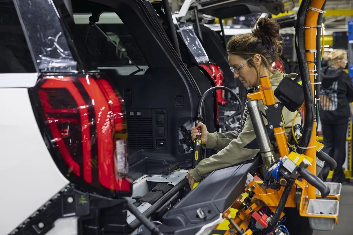 Ford factory worker fitting cargo area trim at the rear of a white SUV body.