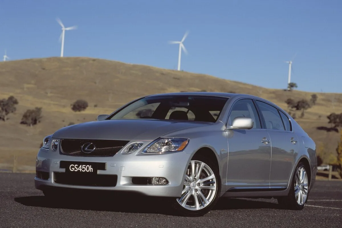 Silver Lexus GS 450h sedan shown from the front left three-quarter angle on a paved road with hills and wind turbines behind.