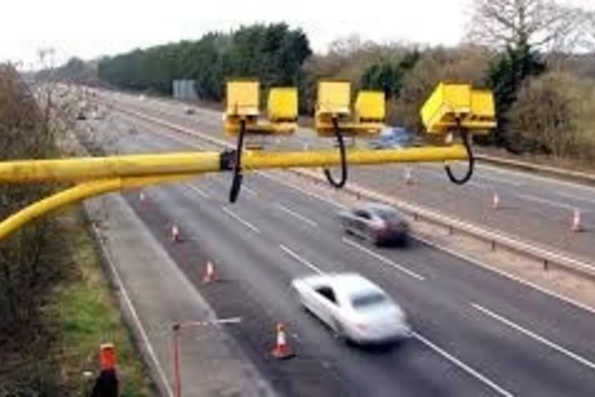 Yellow motorway enforcement gantry above a UK-style multi-lane road with traffic cones and moving cars