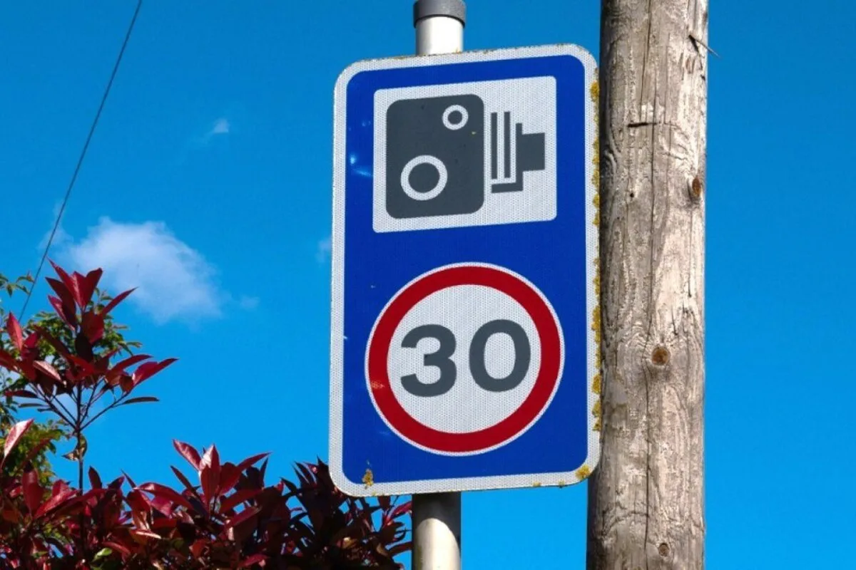 Blue speed camera warning sign above a 30 limit sign beside a wooden pole under a clear sky