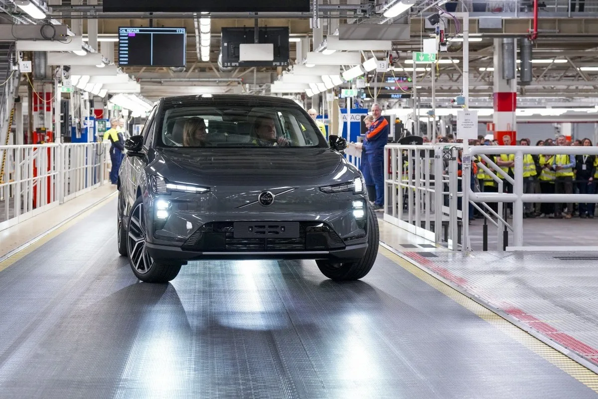 Front view of a grey Volvo EX60 being driven through an indoor assembly plant, with workers watching behind safety barriers.