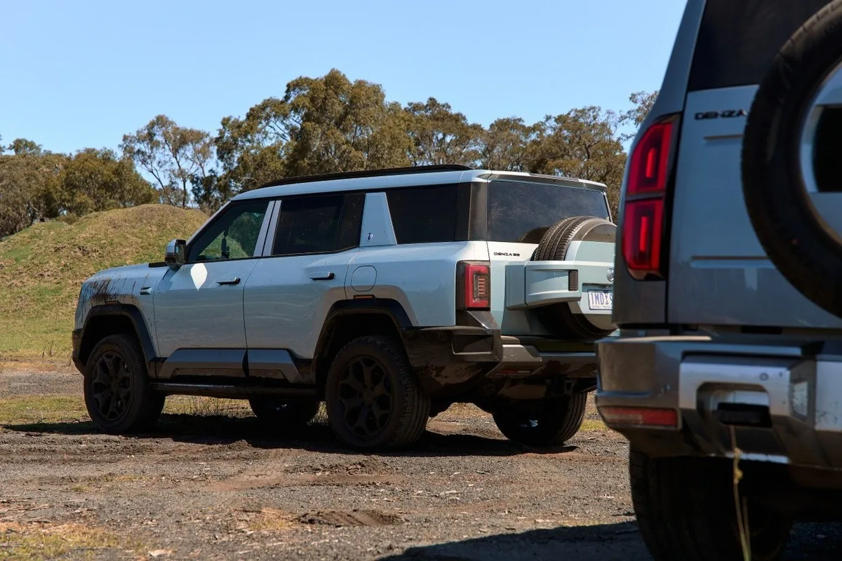 Denza B8 rear three-quarter view on dirt with rear-mounted spare wheel and another Denza B8 in the foreground.