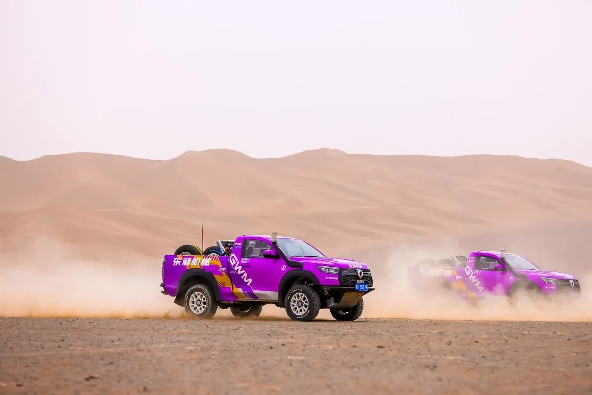 Two magenta GWM TANK rally SUVs crossing a flat desert stage with dust trails behind them.