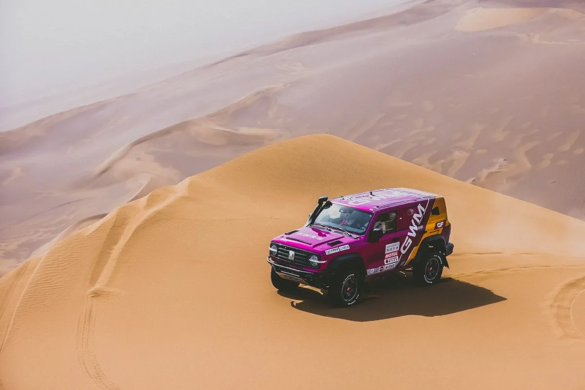 A magenta GWM TANK rally SUV driving alone across wide Taklimakan sand dunes.