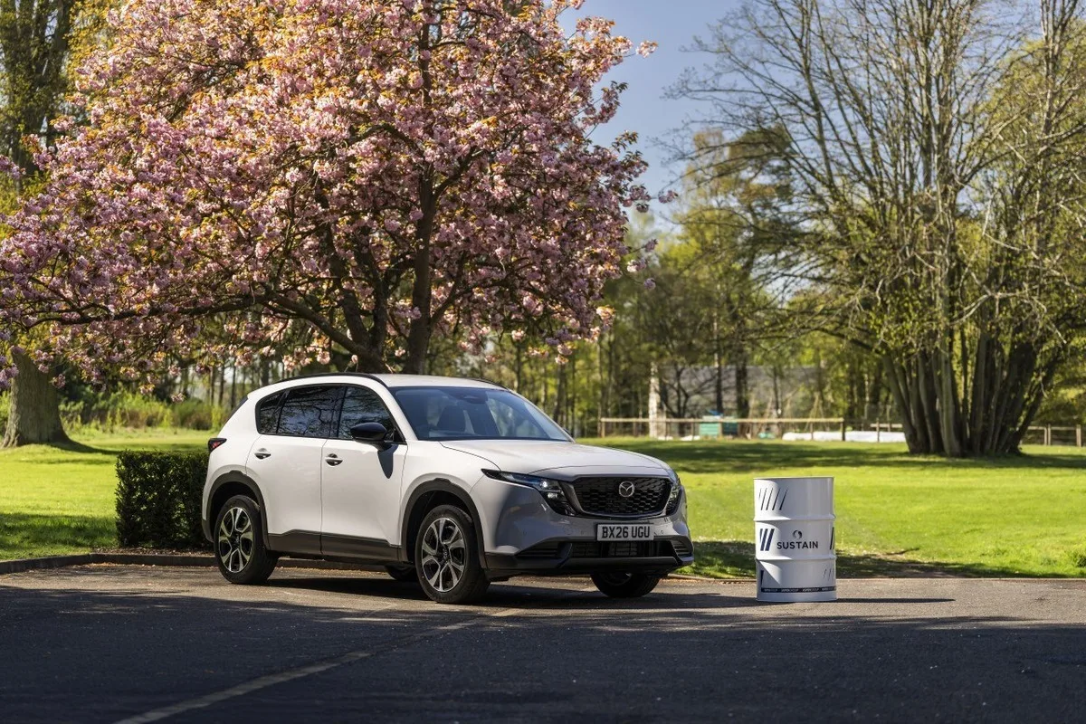 White Mazda CX-5 SUV parked next to a SUSTAIN biofuel drum under a flowering tree
