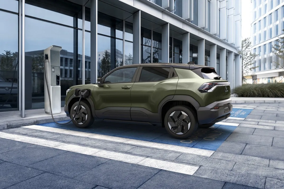 Olive green Suzuki e VITARA parked beside an EV charging station near a modern building.