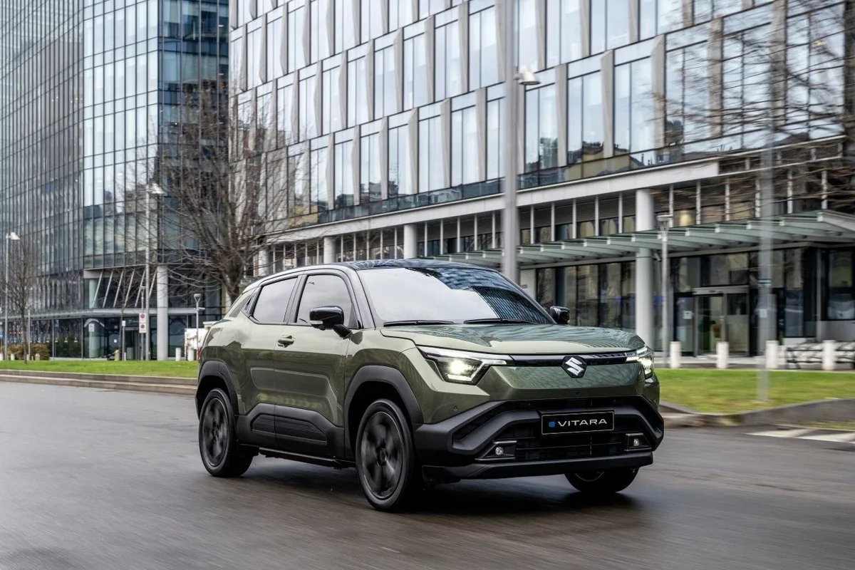 Olive green Suzuki e VITARA driving past glass-fronted city buildings.