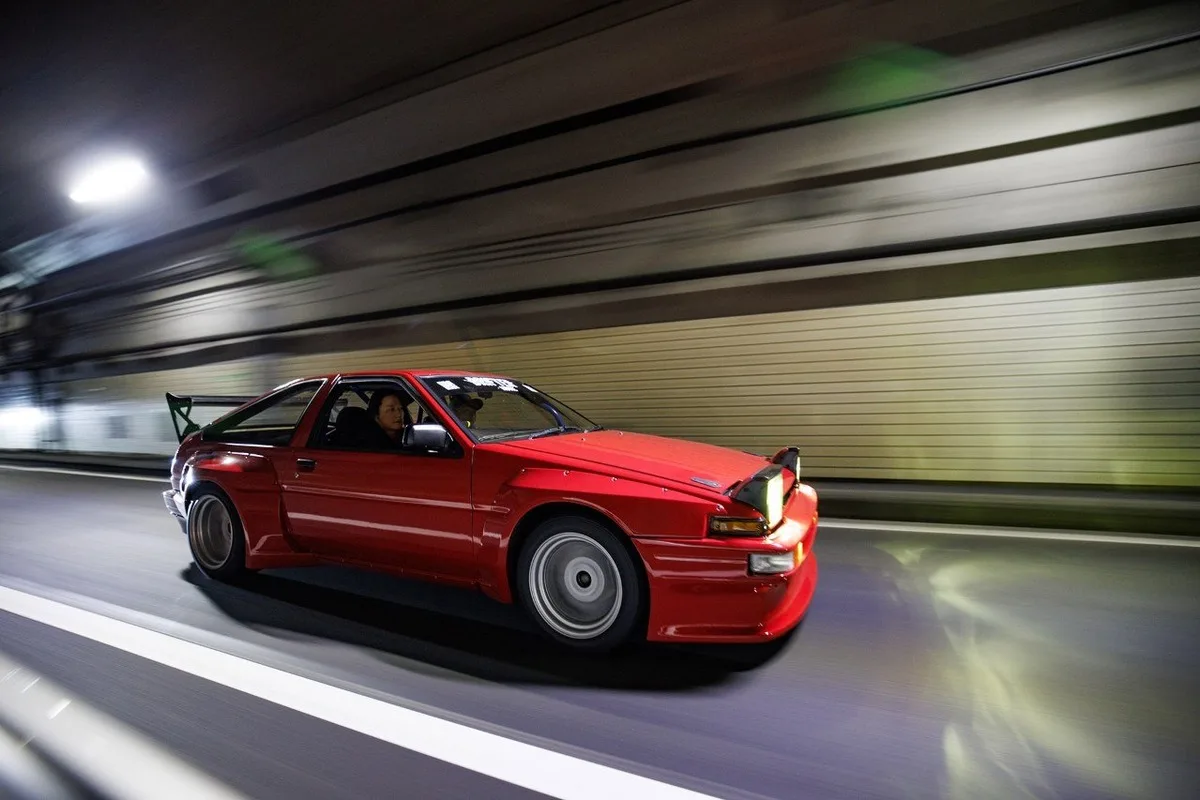 Red modified AE86-style drift car driving through a tunnel at night.