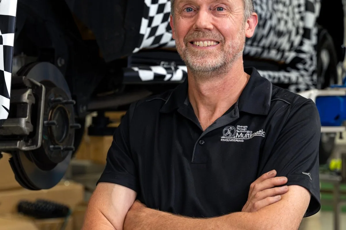 Engineer in a Multimatic polo shirt standing beside a camouflaged JAC Hunter raised on a workshop hoist