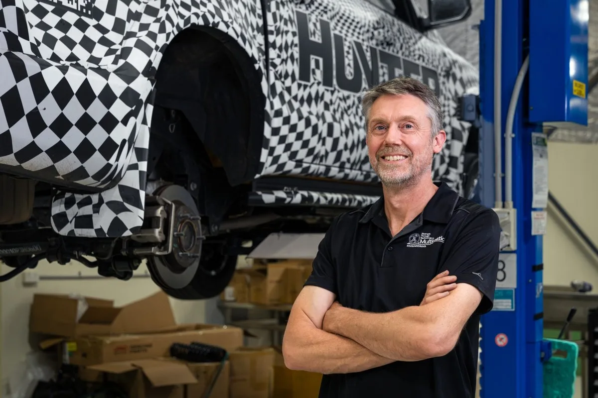 Camouflaged JAC Hunter on a two-post hoist in a workshop with the rear suspension exposed