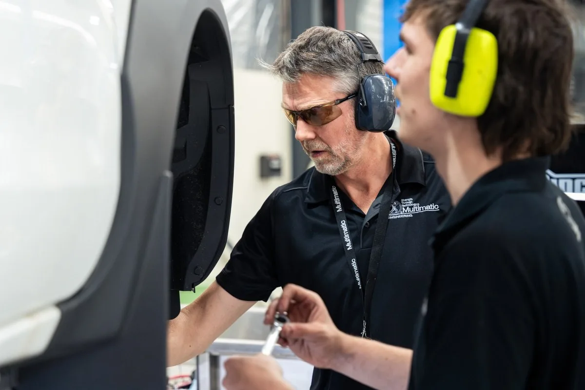 Two engineers wearing ear defenders working at the front wheel arch of a white vehicle in a workshop