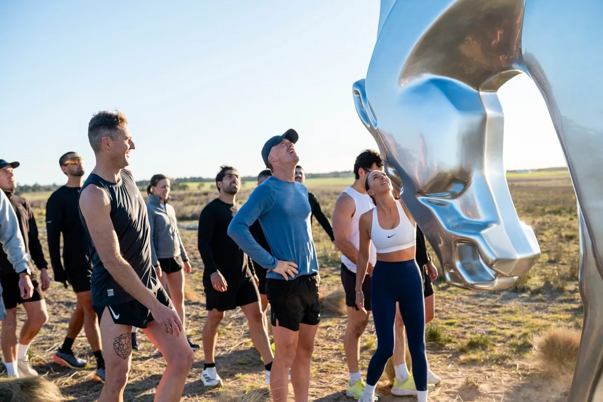 A group of runners in a paddock looking up at the underside of the chrome horse sculpture