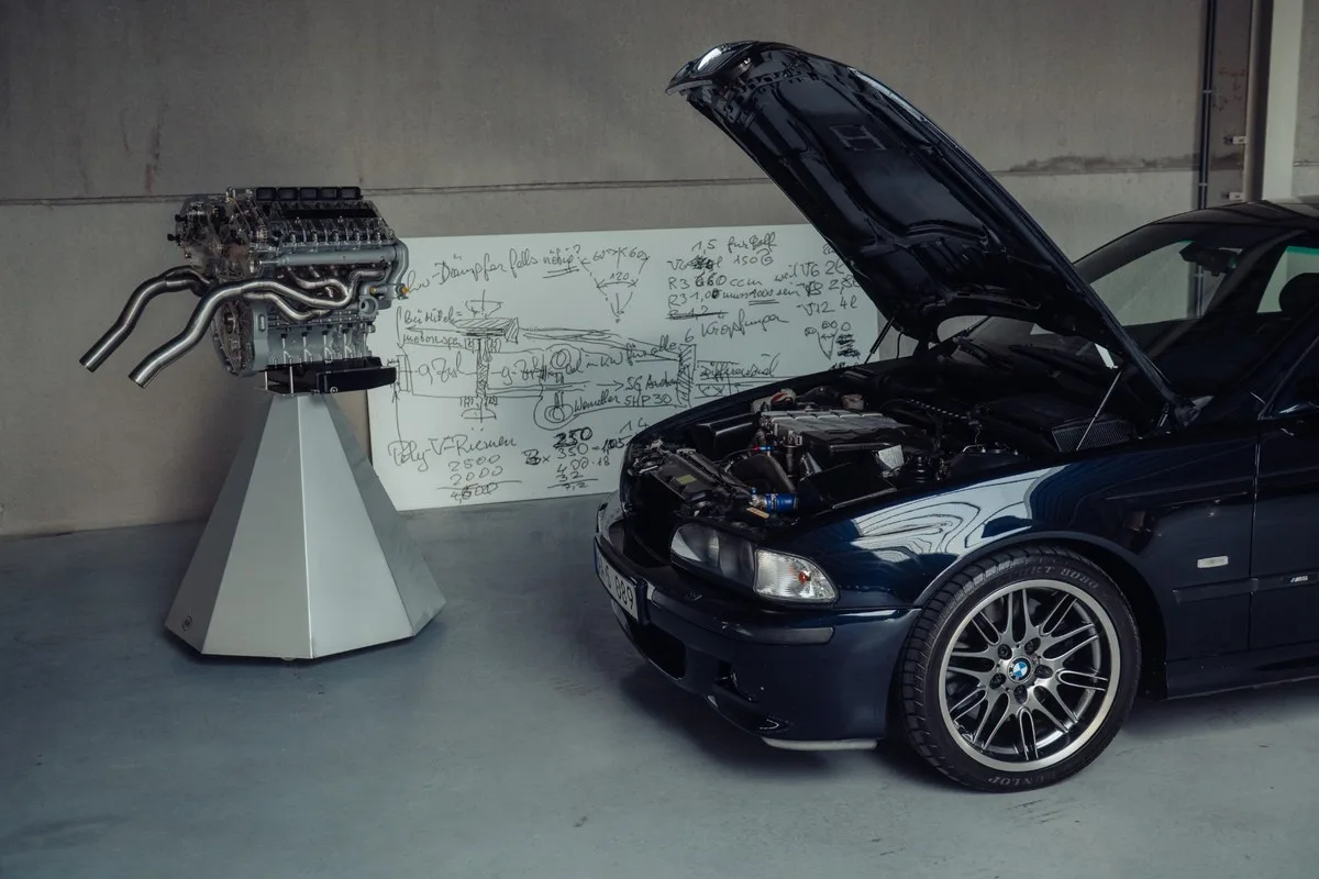 BMW M5 with bonnet open beside a standalone W10 engine and a whiteboard of engineering notes