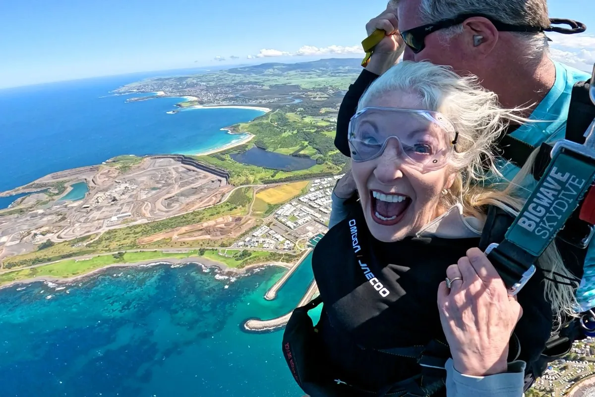 Grey-haired woman in goggles laughing during a tandem skydive over the coast, wearing an Omoda Jaecoo top