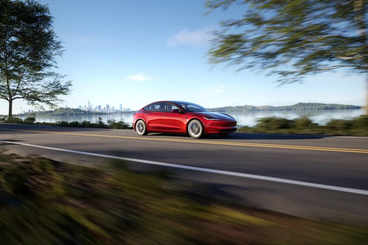 Red Tesla Model 3 driving beside a lake with a city skyline behind